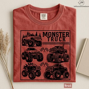 May include: Brick-colored t-shirt featuring a black graphic of four monster trucks with large tires. The text "MONSTER TRUCK" is above the trucks, with checkered flags and trees. The shirt is made of Comfort Colors material.