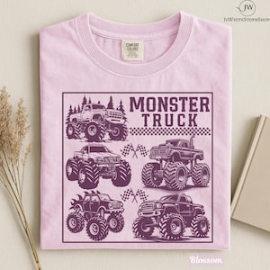 May include: A light pink t-shirt featuring a graphic of several monster trucks in a square frame. The text "MONSTER TRUCK" is displayed above the trucks. The shirt has a tag that says "Comfort Colors".