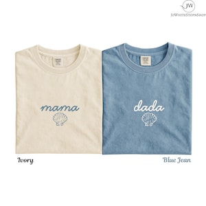 May include: Two short-sleeve crew neck t-shirts. The left shirt is ivory with the word mama and a seashell graphic in blue. The right shirt is blue jean with the word dada and a white seashell graphic. Both feature Comfort Colors neck labels.