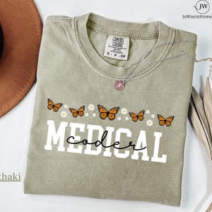 May include: Khaki-coloured t-shirt with the words "MEDICAL coder" in white block letters and a cursive font. The shirt features orange butterflies and white daisies. A silver necklace with a pendant is visible.