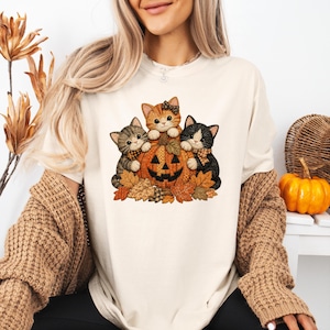 Könnte beinhalten: Ein cremefarbenes T-Shirt mit einem Grafikdruck von drei Kätzchen, die um einen geschnitzten Halloween-Kürbis sitzen. Die Kätzchen sind orange getigert, grau getigert sowie schwarz-weiß und von Herbstblättern in Braun- und Orangetönen umgeben.