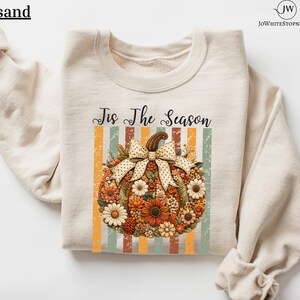 May include: A sand-coloured crewneck sweatshirt featuring a graphic of a pumpkin adorned with various flowers and a polka-dot bow. Above the pumpkin, the text reads 'Tis The Season' in black script, set against a background of vertical stripes.