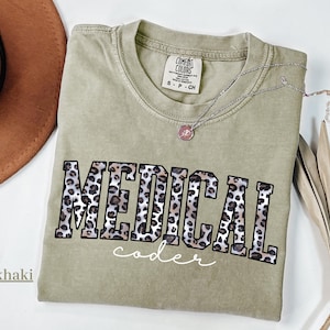May include: Khaki-colored t-shirt with the words "MEDICAL coder" in a leopard print design. The shirt has a crew neck and short sleeves. A silver necklace with a small pendant is also visible.