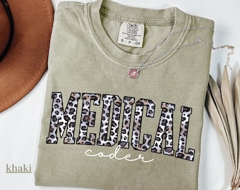 May include: Khaki-colored t-shirt with the words "MEDICAL coder" in a leopard print design. The shirt has a crew neck and short sleeves. A silver necklace with a small pendant is also visible.