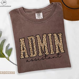 Può includere: Una t-shirt a maniche corte marrone con scollo rotondo. La parte anteriore presenta la parola ADMIN in grandi lettere stampatello con un motivo a giraffa beige e marrone, e la parola assistant scritta sotto in un carattere corsivo nero.