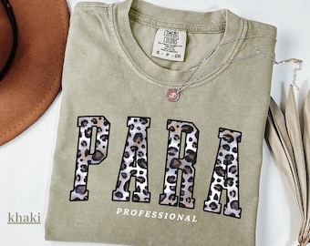 May include: Khaki-colored t-shirt with the word "PARA" in leopard print letters and the word "PROFESSIONAL" underneath. A silver necklace with a small pendant is on the shirt. The shirt is a Comfort Colors brand, size S-P-CH.
