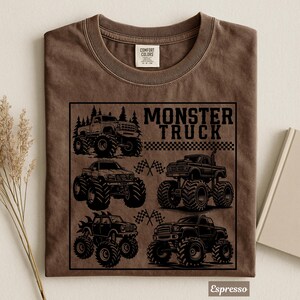 May include: Espresso-colored t-shirt featuring a black graphic of four monster trucks with large tires. The text "MONSTER TRUCK" is above a checkered flag design. The shirt is made of Comfort Colors material.