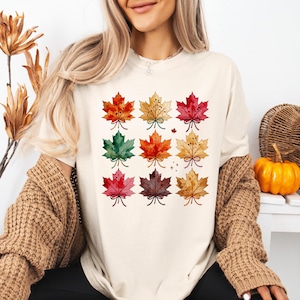 May include: A cream-colored t-shirt featuring a grid of nine watercolor-style maple leaves in various autumn colors including red, orange, green, and brown. Each leaf is tied with a small bow at the stem, and the design includes small star accents.