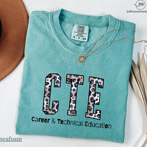 Könnte beinhalten: Türkisfarbenes T-Shirt mit den Buchstaben "CTE" in Leopardenmuster. Darunter steht "Career & Technical Education". Eine goldene Halskette mit Anhänger liegt auf dem Shirt.