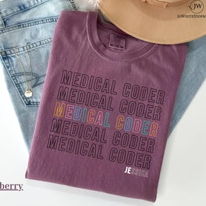 May include: Berry-colored t-shirt with the text "MEDICAL CODER" repeated in a stacked design. The word "CODER" is in multiple colors. The name "JESSICA" is printed at the bottom. The shirt is folded on top of blue jeans.