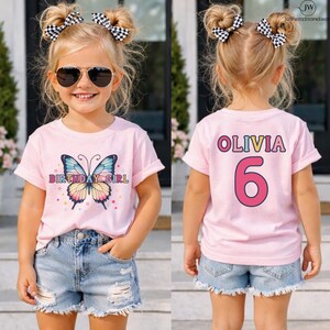 May include: A light pink short-sleeve t-shirt featuring a colorful butterfly graphic with the text "BIRTHDAY GIRL" on the front and the name "OLIVIA" with the number "6" on the back, paired with light blue distressed denim shorts.