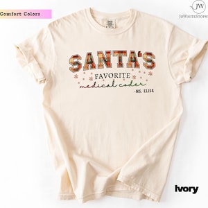 Könnte beinhalten: Elfenbeinfarbenes T-Shirt mit dem Text "SANTA'S FAVORITE medical coder - MS. ELISA" in einem Karomuster. Das Shirt ist von der Marke Comfort Colors und hat einen Rundhalsausschnitt und kurze Ärmel. Der Text ist in einer festlichen Schrift mit Schneeflocken.
