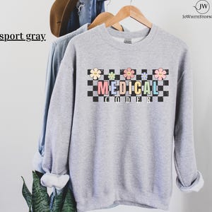Puede incluir: Sudadera gris claro con las palabras "MEDICAL CODER" en letras coloridas, sobre un fondo a cuadros blanco y negro. Encima del texto hay gráficos de flores en colores pastel. Las palabras "sport gray" están en la esquina superior izquierda.
