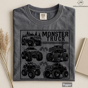 May include: A gray Comfort Colors t-shirt featuring a black graphic of several monster trucks. The text "MONSTER TRUCK" is displayed above the trucks. The shirt is folded and has a tag at the top.