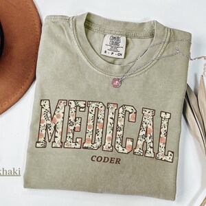 May include: Khaki-coloured t-shirt with the words "MEDICAL CODER" in a floral design. The shirt has a crew neck and short sleeves. A silver necklace with a small pendant is on the shirt.