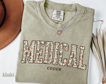 May include: Khaki-colored t-shirt with the words "MEDICAL CODER" in a floral design. The shirt has a crew neck and short sleeves. A silver necklace with a small pendant is on the shirt.