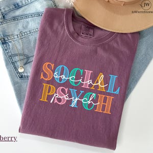 Könnte beinhalten: Ein beerenfarbenes T-Shirt mit den Worten "SOCIAL PSYCH" in farbenfrohem, geschichtetem Text. Das Shirt ist gefaltet und liegt auf einer Jeans, mit einer goldenen Halskette und einem beigen Hut.