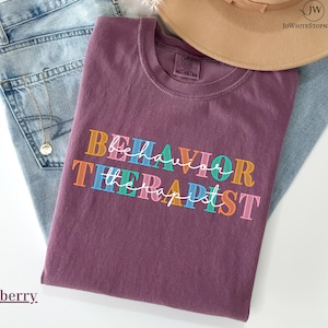 Puede incluir: Camiseta color bayas con las palabras "BEHAVIOR THERAPIST" en letras de bloque coloridas. La camiseta está doblada sobre unos vaqueros azules, con un collar dorado y un sombrero beige en el fondo.