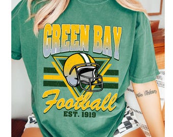 Puede incluir: Camiseta verde con el texto "GREEN BAY Football" y un gráfico de casco de fútbol americano. El diseño utiliza colores amarillo, verde y blanco, y el texto "EST. 1919". La camiseta tiene un aspecto vintage y desgastado.