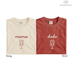 Puede incluir: Dos camisetas Comfort Colors, una marfil y otra color ladrillo. La camiseta marfil tiene la palabra "mama" y un gráfico de langosta en granate. La camiseta color ladrillo tiene la palabra "dada" y un gráfico de langosta en blanco. Las camisetas están etiquetadas como "Ivory" y "Brick".