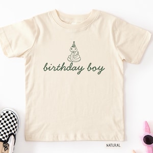 May include: A natural-colored short-sleeve t-shirt featuring a dark green graphic of a coiled snake wearing a party hat. Below the snake, the text "birthday boy" is printed in a dark green cursive font. The shirt is displayed on a plain background.