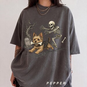 May include: A charcoal grey short-sleeve t-shirt featuring a graphic print of a small brown terrier dog holding a bone in its mouth, chased by a human skeleton near a tombstone marked RIP. The shirt has a crew neckline and a relaxed fit.