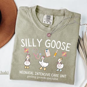 May include: Khaki-colored t-shirt with the words "SILLY GOOSE" and illustrations of cartoon geese wearing medical accessories. The shirt also features the text "NEONATAL INTENSIVE CARE UNIT gosling growth specialist."