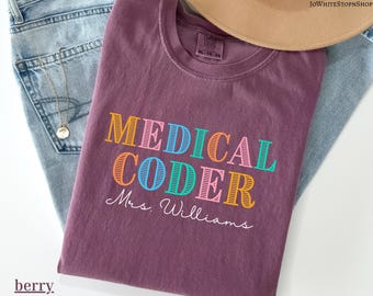 May include: Berry-colored t-shirt with the words "MEDICAL CODER" in colorful block letters and "Mrs. Williams" in script. The shirt is folded on top of blue jeans, with a gold necklace and a tan hat in the background.