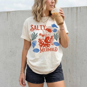 May include: Cream-colored t-shirt with a red and blue graphic. The graphic features the words "Salty Mermaid" with a mermaid illustration, coral, and seashell accents. The shirt has rolled-up sleeves.