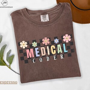 Puede incluir: Camiseta color espresso con un diseño floral y de tablero de ajedrez. La camiseta presenta las palabras "MEDICAL CODER" en letras coloridas. La camiseta está hecha de un material suave.