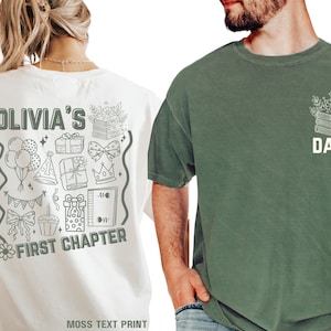 Może przedstawiać: Dwie koszulki, jedna kremowa i jedna zielona. Kremowa koszulka ma napis "OLIVIA'S FIRST CHAPTER" z grafiką urodzinową. Zielona koszulka zawiera słowo "DADA" z motywem książki i kwiatów. Obie to odzież codzienna.