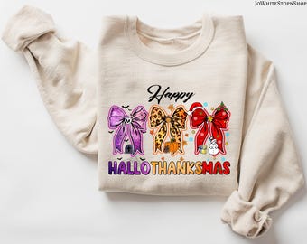 May include: A sand-colored sweatshirt with a graphic that says "Happy HALLOTHANKSMAS." The graphic features three decorative bows: a purple Halloween bow, a leopard print Thanksgiving bow, and a red Christmas bow.