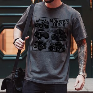 May include: Dark gray t-shirt featuring a black graphic of six monster trucks within a square frame. The text "MONSTER TRUCK" is above the trucks. The shirt has a crew neck and short sleeves.