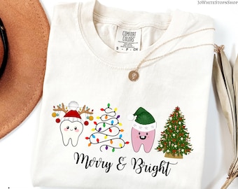 May include: Ivory-colored t-shirt with a Christmas-themed graphic. The design features cartoon teeth wearing Santa hats and decorated as Christmas trees, with the words "Merry & Bright" in a script font.