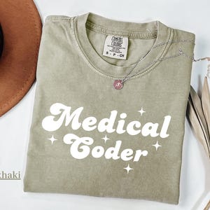 May include: Khaki-colored t-shirt with the words "Medical Coder" in white, retro-style font. The shirt has a crew neck and short sleeves. A silver necklace with a pendant is draped across the shirt.