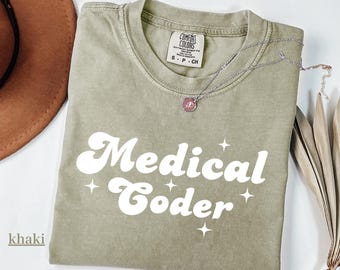 May include: Khaki-colored t-shirt with the words "Medical Coder" in white, retro-style font. The shirt has a crew neck and short sleeves. A silver necklace with a pendant is draped across the shirt.