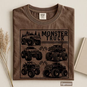 May include: Espresso-colored t-shirt featuring a black graphic of four monster trucks with large tires. The text "MONSTER TRUCK" is above a checkered flag design. The shirt is made of Comfort Colors material.