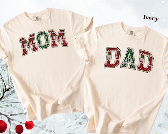 May include: Two ivory-colored t-shirts with the words "MOM" and "DAD" printed in a festive, collegiate-style font. The letters are outlined in red and filled with a green and red pattern. The shirts are displayed on a light background.