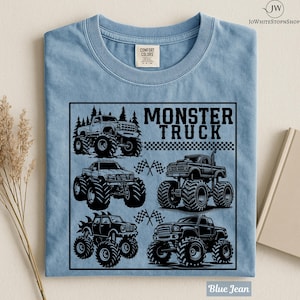 May include: Blue jean colored t-shirt featuring a black graphic of several monster trucks with large tires. The words "MONSTER TRUCK" are printed in black, with a checkered flag design. The shirt is made of a soft, comfortable material.