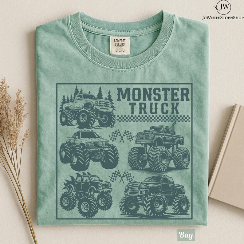 May include: A light green t-shirt featuring a graphic of several monster trucks in a square frame. The words "MONSTER TRUCK" are printed above the trucks. The shirt is made of a soft material and has a crew neck.