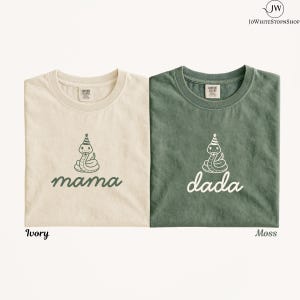 Peut inclure: Deux t-shirts à col rond, un ivoire et un vert mousse, chacun arborant un motif de serpent portant un chapeau de fête. Le t-shirt ivoire porte l'inscription mama en vert, tandis que le t-shirt vert mousse porte l'inscription dada en blanc.