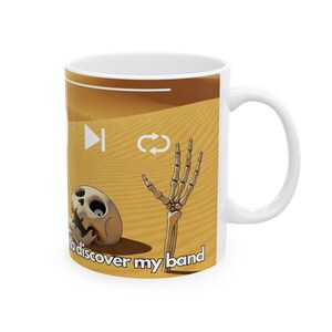 Könnte beinhalten: Weiße Keramiktasse mit einer Wüstenlandschaft, die eine Skeletthand zeigt, die aus dem Sand ragt, und einen teilweise vergrabenen Schädel. Die Tasse hat einen weißen Hintergrund mit einer braunen Wüstenlandschaft und dem Text "to discover my band".