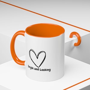 Op de afbeelding: Witte keramische mok met een oranje rand en handvat. De mok heeft een zwart handgetekend hart en de tekst "Single and Looking" gedrukt op de voorkant.