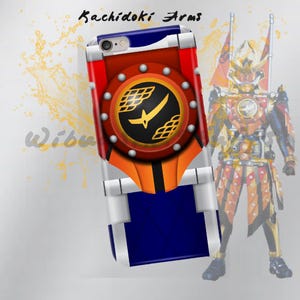 May include: A phone case with a red, orange, blue, and silver design. The case features the text "Kachidoki Arms" and a circular emblem with a yellow sword and geometric shapes. A partially visible character is in the background.