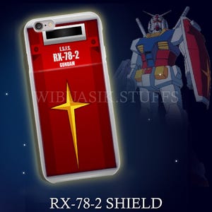 May include: A phone case with a red shield design, a yellow star, and the text "RX-78-2 GUNDAM." The case has a white border and a black rectangle at the top. A Gundam robot is in the background.