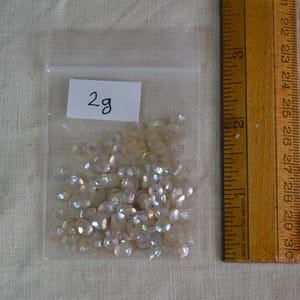 May include: A small clear plastic bag containing iridescent sequins, labelled "2g". A wooden ruler is positioned next to the bag. The sequins appear to be a light, pearlescent colour.