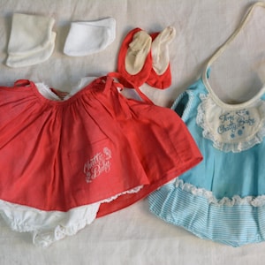 May include: A set of doll clothing including a red dress with white lace trim and the text Chatty Baby, a light blue romper with white lace and striped trim featuring the text Tiny Chatty Baby, two white socks, and two small red shoes.
