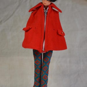 May include: A vintage doll wearing a bright red coat with a silver zipper and two small pockets. The doll has black hair and is wearing patterned pants with red, blue, and green geometric designs. The doll is standing on a white stand.