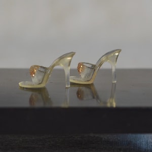 May include: A pair of clear, high-heeled shoes with a glittery gold accent on the toe strap. The heels are slender and transparent, and the shoes are set against a dark surface. The overall design is delicate and elegant.