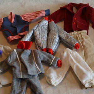May include: A collection of clothing items for a doll, including a red and white striped jacket, a silver jacket, a grey jacket, a white shirt, a pair of beige trousers, and a white shirt with red trim.